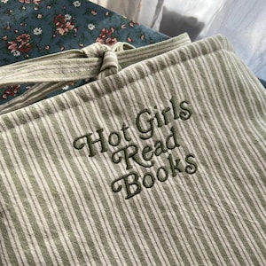 Puede incluir: Una funda para libros a rayas verdes y blancas con el texto bordado "Hot Girls Read Books" en hilo verde oscuro. La funda tiene un cierre de lazo y est&aacute; hecha de una tela suave y texturizada. El fondo incluye tela floral.