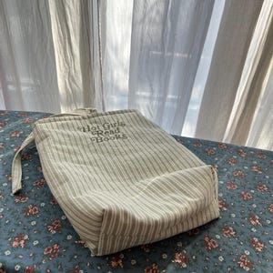Puede incluir: Una bolsa con cord&oacute;n beige y blanco a rayas con el texto "Hot Girls Read Books". La bolsa est&aacute; sobre una superficie azul con estampado floral. La bolsa tiene un asa de lazo.