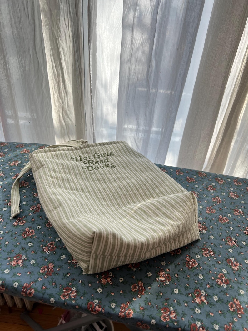 Puede incluir: Una bolsa con cord&oacute;n beige y blanco a rayas con el texto "Hot Girls Read Books". La bolsa est&aacute; sobre una superficie azul con estampado floral. La bolsa tiene un asa de lazo.