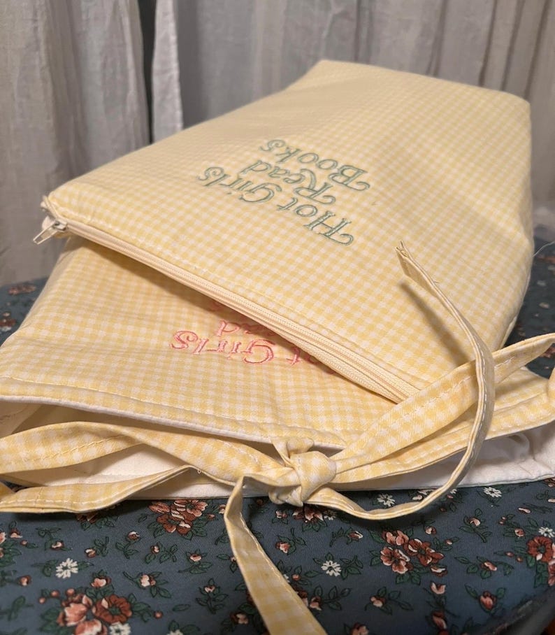 Puede incluir: Dos fundas de libros de cuadros amarillos con texto bordado. Una funda dice "Hot Girls Read Books" en hilo verde, la otra "Girls" en hilo rosa. Las fundas est&aacute;n atadas con una cinta amarilla.