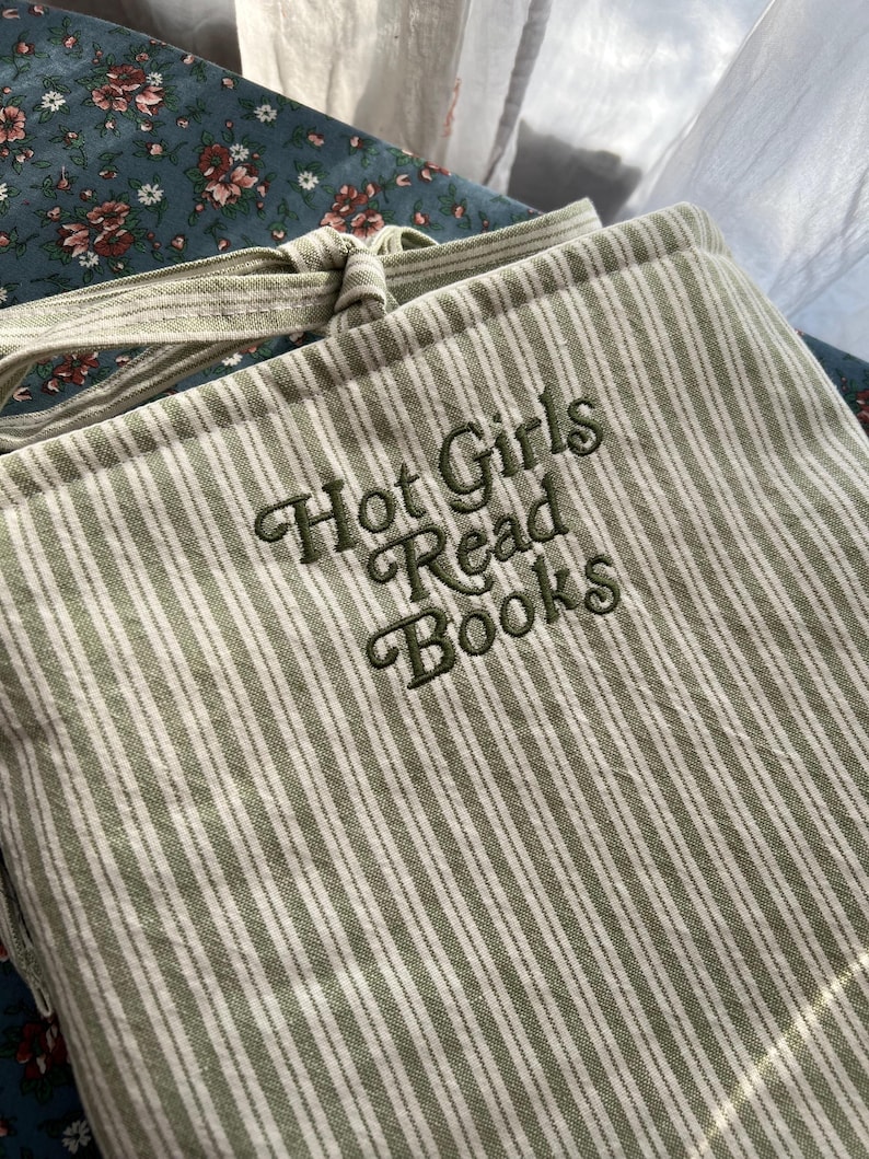 Puede incluir: Una funda para libros a rayas verdes y blancas con el texto bordado "Hot Girls Read Books" en hilo verde oscuro. La funda tiene un cierre de lazo y est&aacute; hecha de una tela suave y texturizada. El fondo incluye tela floral.