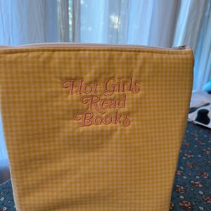 Puede incluir: Una bolsa con cremallera de cuadros amarillos con el texto bordado "Hot Girls Read Books" en una fuente cursiva. La bolsa es rectangular y parece estar hecha de tela.
