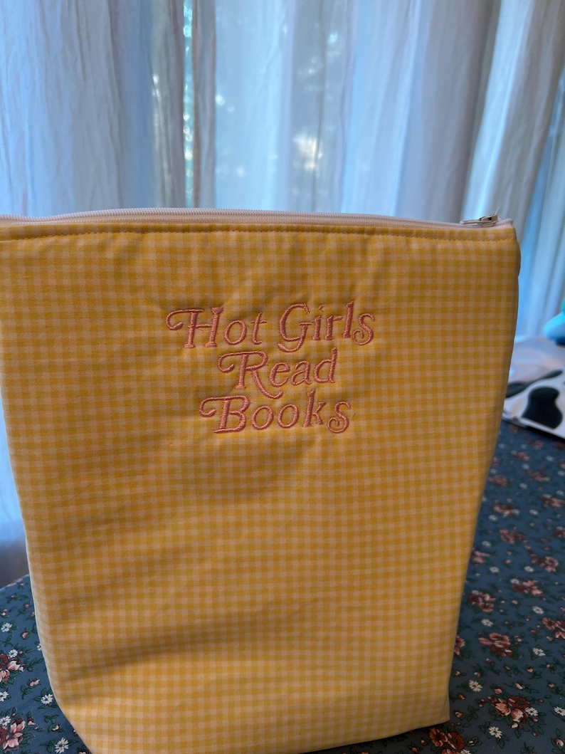 Puede incluir: Una bolsa con cremallera de cuadros amarillos con el texto bordado "Hot Girls Read Books" en una fuente cursiva. La bolsa es rectangular y parece estar hecha de tela.
