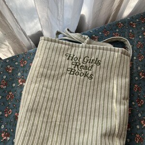 Puede incluir: Un bolso tote de lona con rayas verticales en verde claro y blanco. El bolso tiene una correa para el hombro y el texto bordado "Hot Girls Read Books" en una fuente de escritura. El fondo es una tela estampada floral.