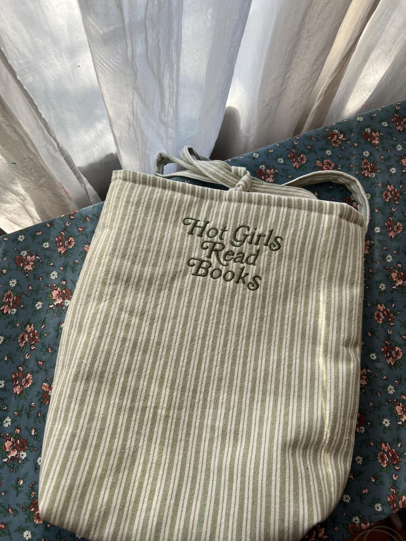Puede incluir: Un bolso tote de lona con rayas verticales en verde claro y blanco. El bolso tiene una correa para el hombro y el texto bordado "Hot Girls Read Books" en una fuente de escritura. El fondo es una tela estampada floral.