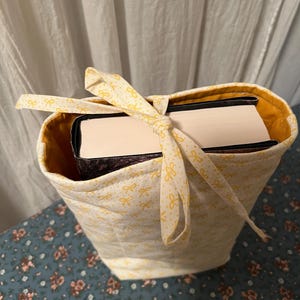 May include: A quilted book tote in a light yellow fabric with a repeating pattern of small yellow bows. The interior is lined with a solid yellow fabric. A matching ribbon ties the top closed, holding a book inside.
