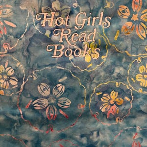 Puede incluir: Una tela turquesa con un estampado floral y el texto bordado "Hot Girls Read Books" en una fuente cursiva. Las flores son en tonos de blanco, amarillo y rosa, con un efecto acuarela.