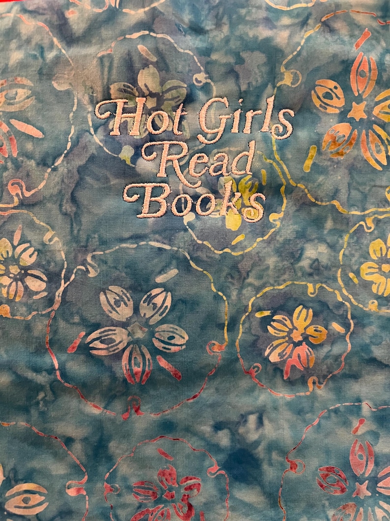 Puede incluir: Una tela turquesa con un estampado floral y el texto bordado "Hot Girls Read Books" en una fuente cursiva. Las flores son en tonos de blanco, amarillo y rosa, con un efecto acuarela.
