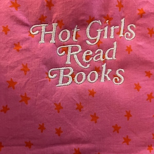 Puede incluir: Tela rosa con patrones de estrellas naranjas y el texto bordado "Hot Girls Read Books" en escritura blanca. La tela tiene una textura ligeramente arrugada.