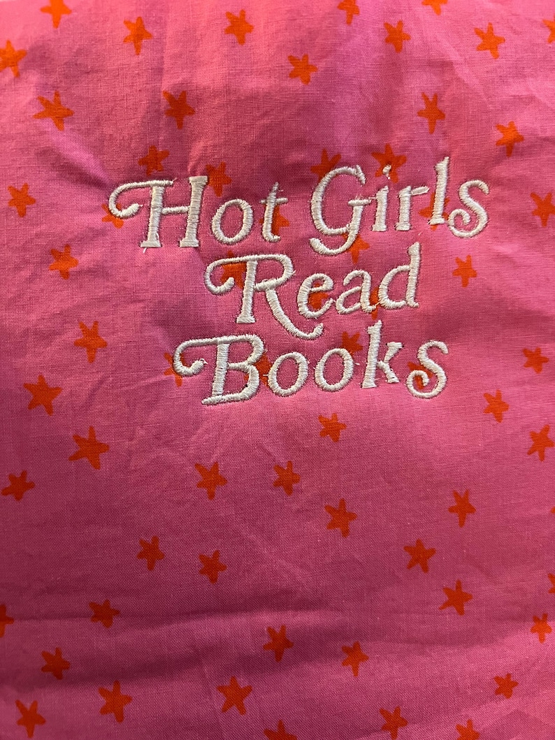 Puede incluir: Tela rosa con patrones de estrellas naranjas y el texto bordado "Hot Girls Read Books" en escritura blanca. La tela tiene una textura ligeramente arrugada.