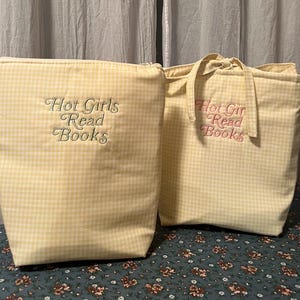 Puede incluir: Dos bolsas de tela de cuadros amarillos. Una es una bolsa con cremallera, la otra es un bolso de mano. Ambas tienen el texto bordado "Hot Girls Read Books". Las bolsas est&aacute;n sobre una superficie con estampado floral.
