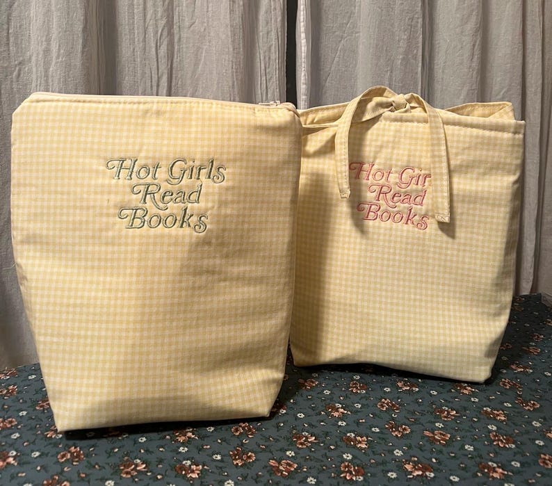 Puede incluir: Dos bolsas de tela de cuadros amarillos. Una es una bolsa con cremallera, la otra es un bolso de mano. Ambas tienen el texto bordado "Hot Girls Read Books". Las bolsas est&aacute;n sobre una superficie con estampado floral.