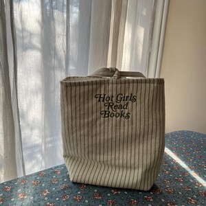 Puede incluir: Un bolso tote a rayas beige y blancas con el texto bordado "Hot Girls Read Books". El bolso tiene forma rectangular con un asa en la parte superior. El bolso est&aacute; sobre una superficie con estampado floral.