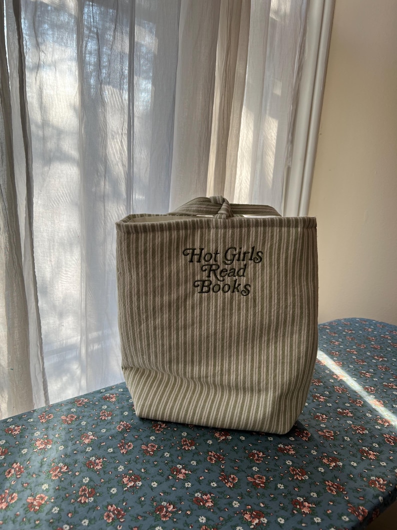 Puede incluir: Un bolso tote a rayas beige y blancas con el texto bordado "Hot Girls Read Books". El bolso tiene forma rectangular con un asa en la parte superior. El bolso est&aacute; sobre una superficie con estampado floral.