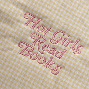 Puede incluir: Primer plano de una tela de cuadros amarillos y blancos con texto rosa bordado que dice "Hot Girls Read Books". El bordado tiene una fuente cursiva delicada.