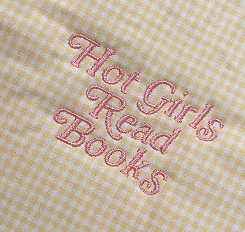 Puede incluir: Primer plano de una tela de cuadros amarillos y blancos con texto rosa bordado que dice "Hot Girls Read Books". El bordado tiene una fuente cursiva delicada.