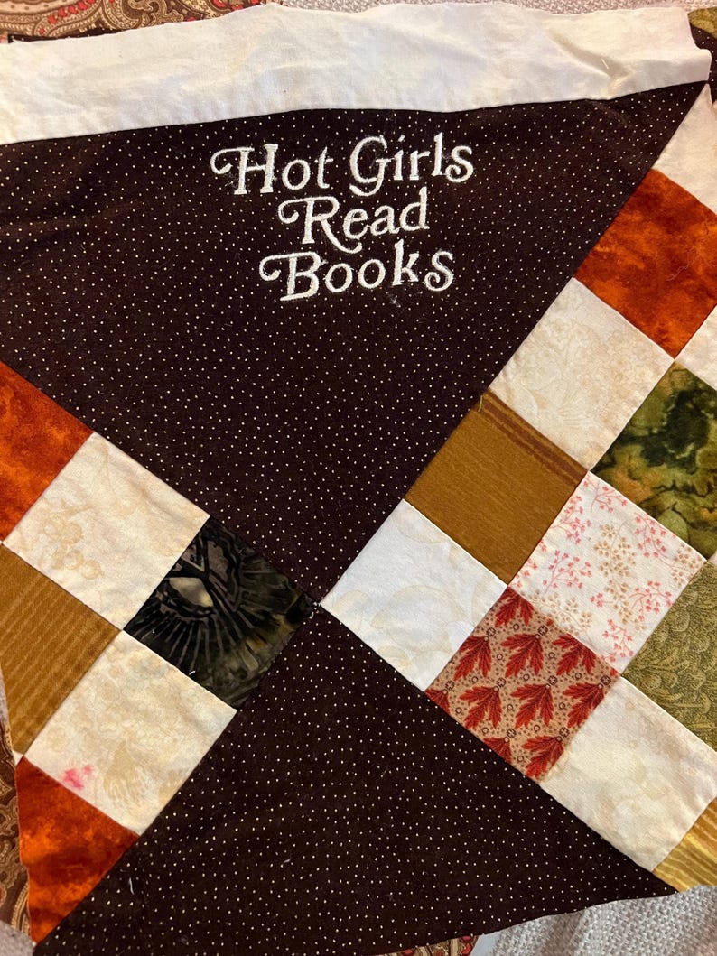 Puede incluir: Un edred&oacute;n hecho a mano con una secci&oacute;n marr&oacute;n con lunares bordada con las palabras "Hot Girls Read Books" en escritura blanca. El edred&oacute;n presenta un dise&ntilde;o de patchwork con cuadrados de varios colores y patrones, incluyendo estampados naranjas, blancos y florales.