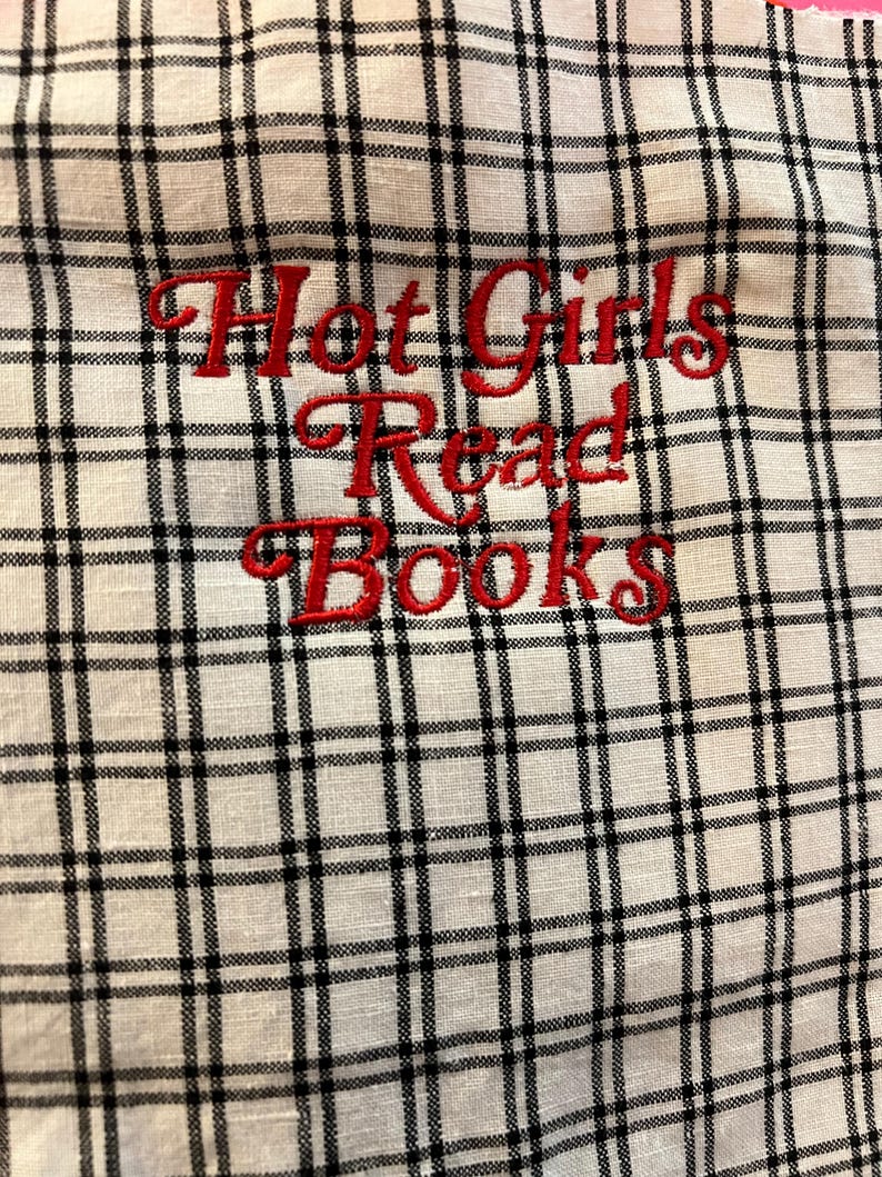 Puede incluir: Tela blanca con un patr&oacute;n de cuadros negros. Las palabras "Hot Girls Read Books" est&aacute;n bordadas en rojo. La tela tiene un aspecto ligeramente texturizado.