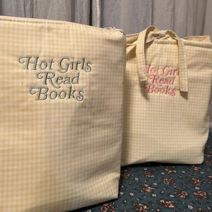 Puede incluir: Dos bolsas de tela de cuadros amarillos. Una es un estuche con cremallera con el texto bordado "Hot Girls Read Books" en verde. La otra es una bolsa de mano con el mismo texto en rosa. Ambas bolsas tienen un estampado de cuadros amarillos claros y blancos.