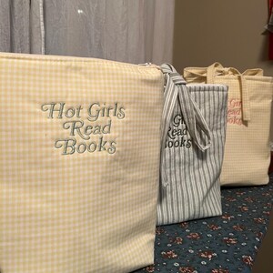 Puede incluir: Tres bolsas de tela con los textos "Hot Girls Read Books" y "Girls Read Books" bordados. Las bolsas son de cuadros amarillos, rayas grises y blancas y un estampado de cuadros amarillo claro. Las bolsas est&aacute;n sobre una superficie con estampado floral.