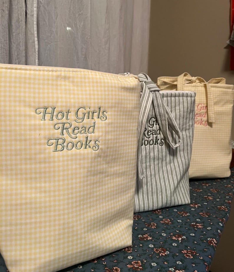 Puede incluir: Tres bolsas de tela con los textos "Hot Girls Read Books" y "Girls Read Books" bordados. Las bolsas son de cuadros amarillos, rayas grises y blancas y un estampado de cuadros amarillo claro. Las bolsas est&aacute;n sobre una superficie con estampado floral.