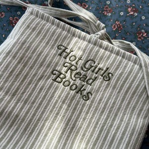 Puede incluir: Un bolso tote a rayas verdes y blancas con el texto bordado "Hot Girls Read Books" en cursiva. El bolso tiene correas largas y est&aacute; hecho de una tela suave. El fondo es un estampado floral.