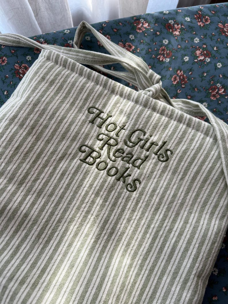 Puede incluir: Un bolso tote a rayas verdes y blancas con el texto bordado "Hot Girls Read Books" en cursiva. El bolso tiene correas largas y est&aacute; hecho de una tela suave. El fondo es un estampado floral.