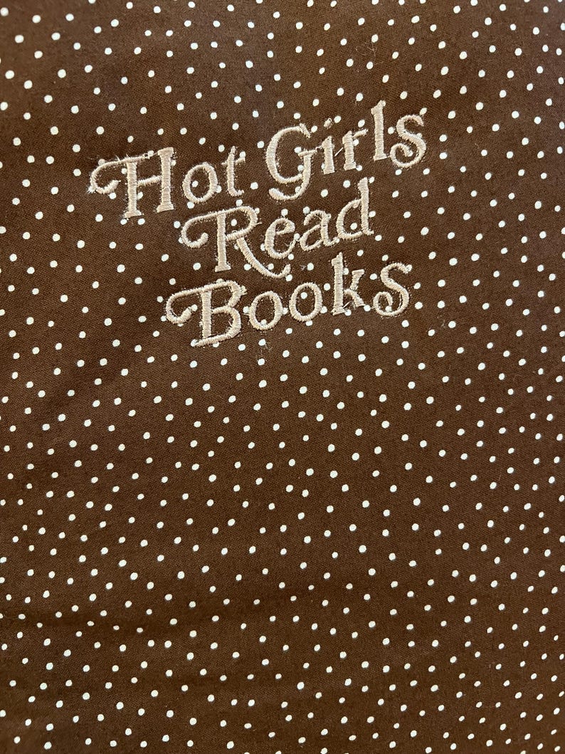 Puede incluir: Tela marr&oacute;n con lunares blancos y texto bordado que dice "Hot Girls Read Books". El texto est&aacute; en una fuente cursiva. La tela es de color marr&oacute;n liso.