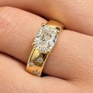 May include: A gold ring featuring a large, rectangular cushion-cut center stone held by four prongs. The wide, polished gold band is decorated with three small, star-shaped engravings, each set with a tiny, sparkling white gemstone.