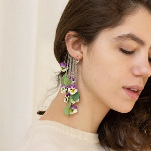 May include: A long, dangling earring with a silver chain and multiple purple and white flowers. The flowers are made of polymer clay and resemble pansies.