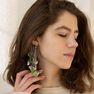 May include: A woman with long brown hair wearing a white sweater and a long, dangling earring with purple and green flowers.