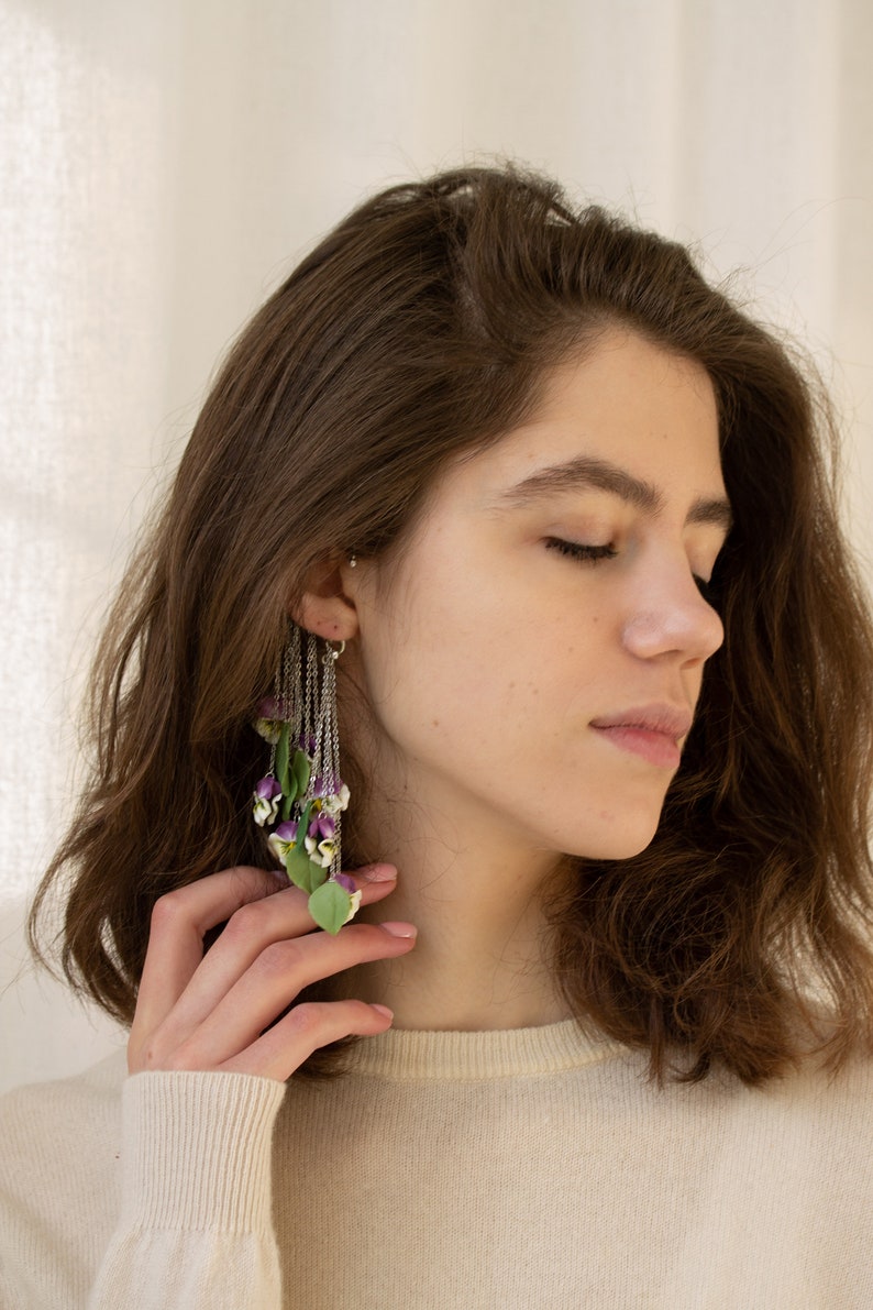 May include: A woman with long brown hair wearing a white sweater and a long, dangling earring with purple and green flowers.