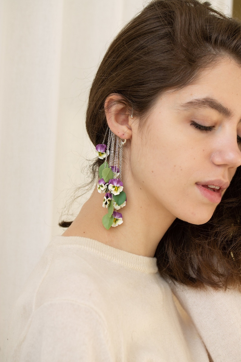 May include: A long, dangling earring with a silver chain and multiple purple and white flowers. The flowers are made of polymer clay and resemble pansies.
