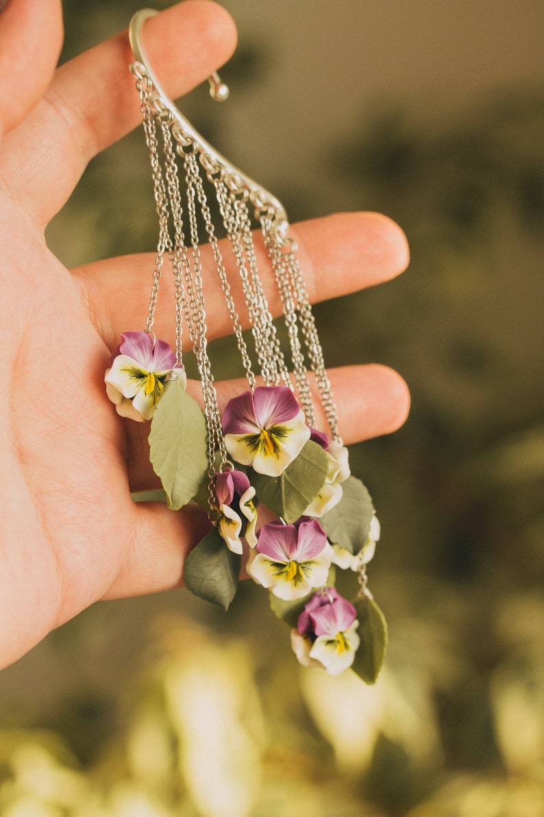 May include: A silver ear cuff with multiple chains and purple and white flower charms. The flowers are made of polymer clay and resemble pansies.