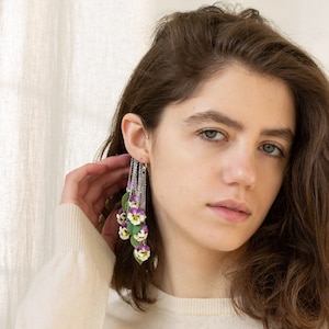 May include: A woman wearing a white sweater with a long, dangling earring featuring a cluster of purple and white flowers. The earring is made of silver chain and has a delicate floral design.