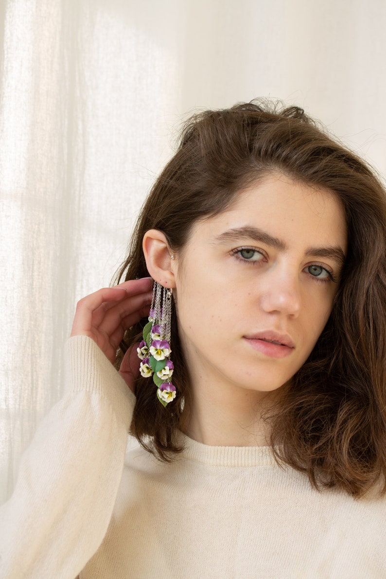 May include: A woman wearing a white sweater with a long, dangling earring featuring a cluster of purple and white flowers. The earring is made of silver chain and has a delicate floral design.
