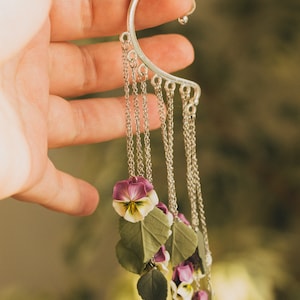 May include: Silver ear cuff with a chain and multiple purple and white flower charms. The flowers are made of polymer clay and resemble pansies.