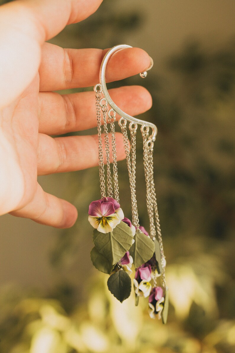 May include: Silver ear cuff with a chain and multiple purple and white flower charms. The flowers are made of polymer clay and resemble pansies.