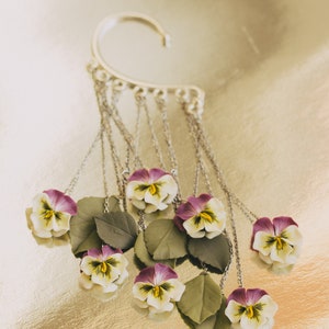 May include: A silver ear cuff with multiple chains hanging down, each chain ending with a small purple and white flower with green leaves.