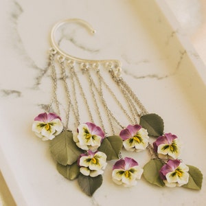 May include: Silver ear cuff with a chain dangling multiple purple and white pansy flowers with green leaves.