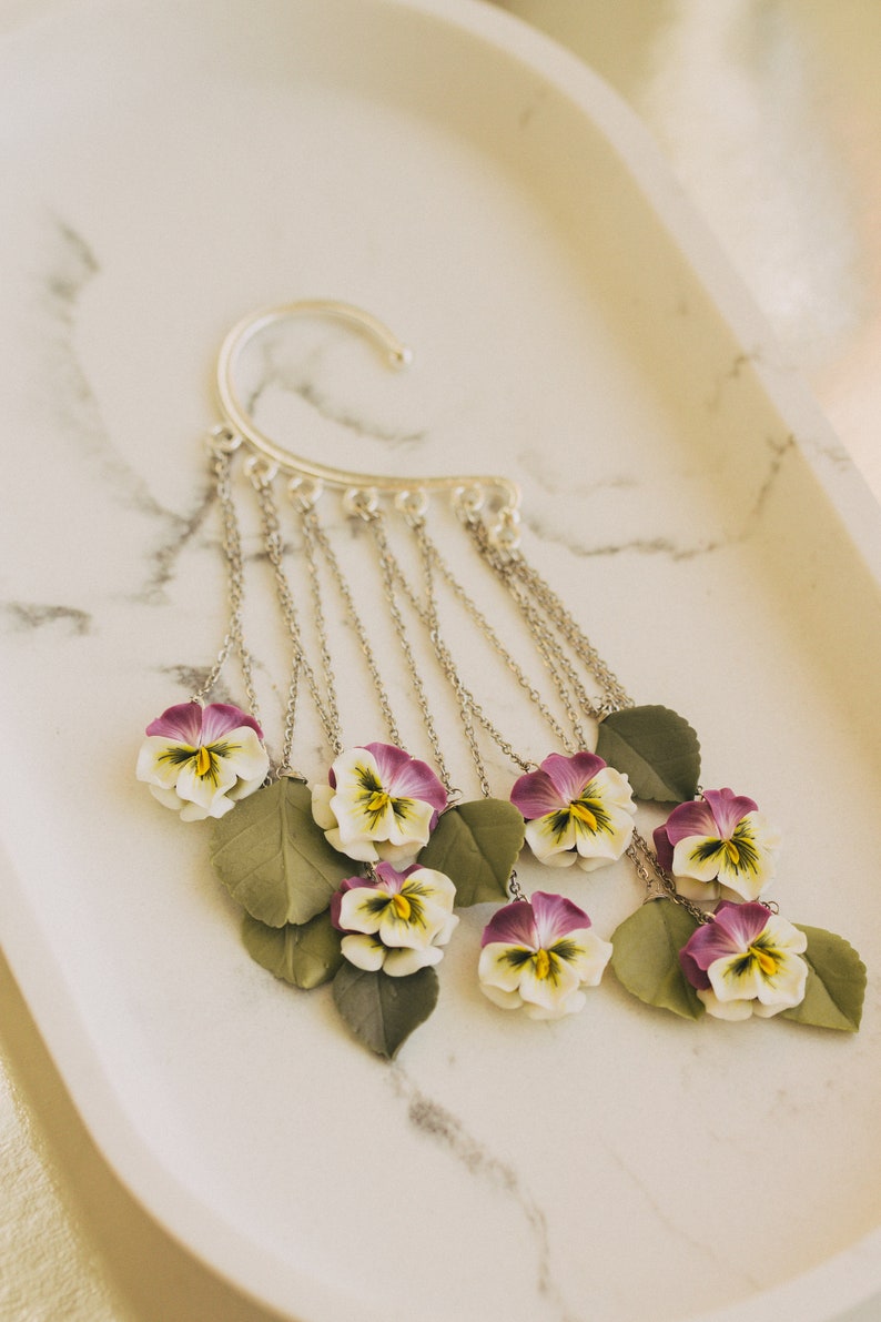 May include: Silver ear cuff with a chain dangling multiple purple and white pansy flowers with green leaves.