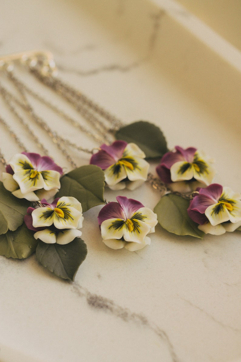 May include: A silver chain necklace with multiple pendants featuring purple and white pansies with green leaves.