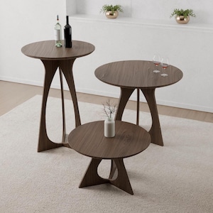 May include: Three round tables with a dark wood grain finish and unique arched bases. The tallest table holds two wine bottles, the medium table holds two wine glasses, and the shortest table features a small white vase with a decorative twig.