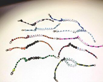 Könnte beinhalten: Eine Sammlung von Armbändern mit Perlen, auf denen verschiedene Teamnamen mit weißen Buchstabenperlen geschrieben sind. Die Armbänder haben verschiedene Farben, darunter Schwarz, Rot, Blau, Grün und Orange, und sind mit silbernen Verschlüssen versehen.