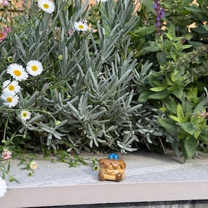 May include: A miniature ceramic pancake figure with a smiling face, brown syrup, and blue blueberry details. The small food sculpture is set against a backdrop of greenery and flowers, including daisies and lavender.