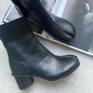 Peut inclure: Paire de bottines en cuir noir avec un design unique à bout fendu. Les bottines ont un petit talon bloc et une finition lisse et polie. Les chaussures sont présentées sur un magazine ouvert avec des photos en noir et blanc.