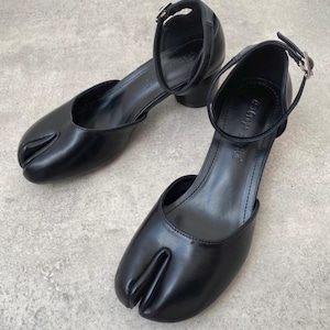 May include: A pair of black leather Tabi shoes with a split-toe design. The shoes have a low heel, an ankle strap with a silver buckle, and a rounded toe. The shoes are on a grey surface.