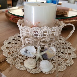 May include: A crocheted candle holder with a white candle and seashells. The holder is decorated with a brown twine bow and is sitting on a crocheted doily.