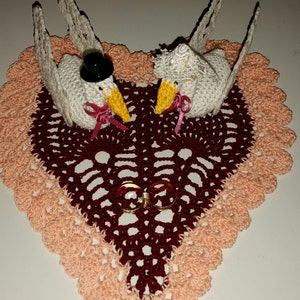 Puede incluir: Un portaanillos de boda hecho a mano con ganchillo con dos cisnes blancos con picos amarillos y lazos rosas, sobre un tapete en forma de corazón burdeos. El tapete está bordeado con ganchillo color melocotón y contiene dos anillos de oro.