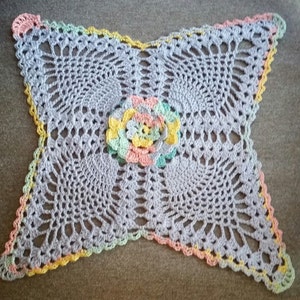 Könnte beinhalten: Eine gehäkelte Deckchen mit einem floralen Design in der Mitte. Die Deckchen ist aus einem hellvioletten Garn gefertigt und hat einen Rand aus gelbem, rosa und grünem Garn.